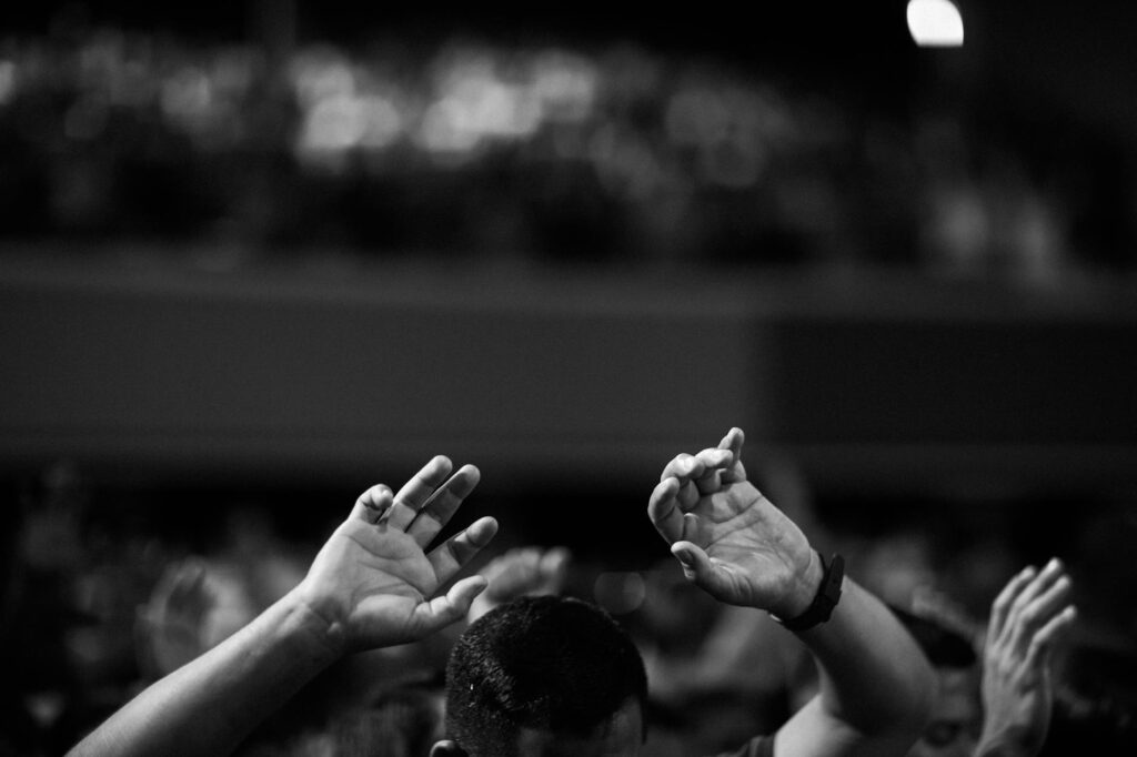 A black and white photo of hands raised in a crowd during a concert or religious gathering indoors.